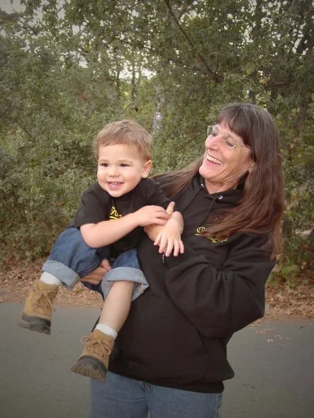 A woman holding a smiling young boy outdoors in a park with trees in the background.