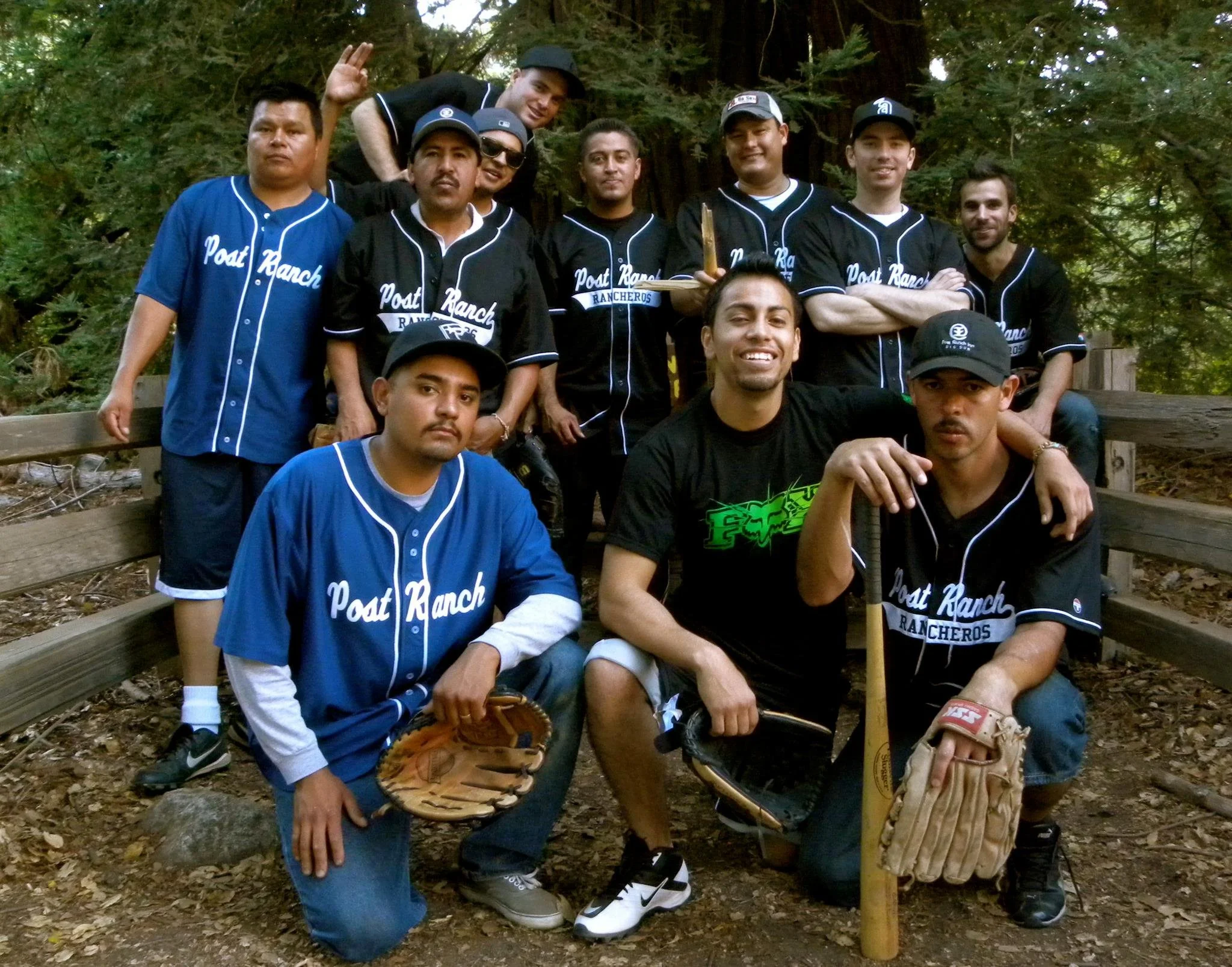 A group of men posing outdoors, wearing baseball jerseys with "Post Ranch Rancheros" printed on them. They are surrounded by trees and foliage, indicating a forest setting. Some are holding baseball gloves, and one has a baseball bat.