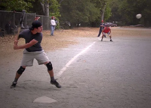 A boy in black shirt and shorts wearing a cap is batting in a baseball game on a dirt field, with a catcher in red uniform and spectators in the background.