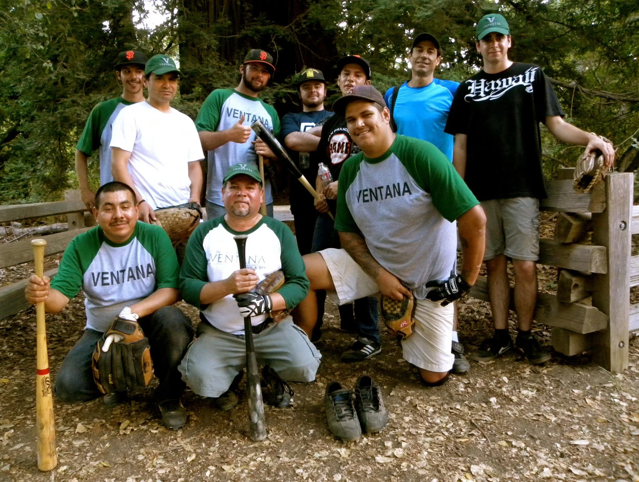 A group of ten men posing outdoors in a wooded area, some are holding baseball bats and wearing casual outdoor clothing, including hats and gloves.