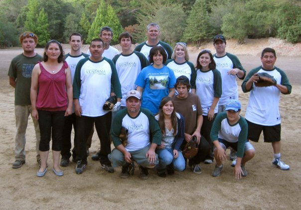 Group of young people and adults outdoors on a sandy area with trees in the background, some wearing matching baseball-style shirts with 'VENYANA' printed on them.