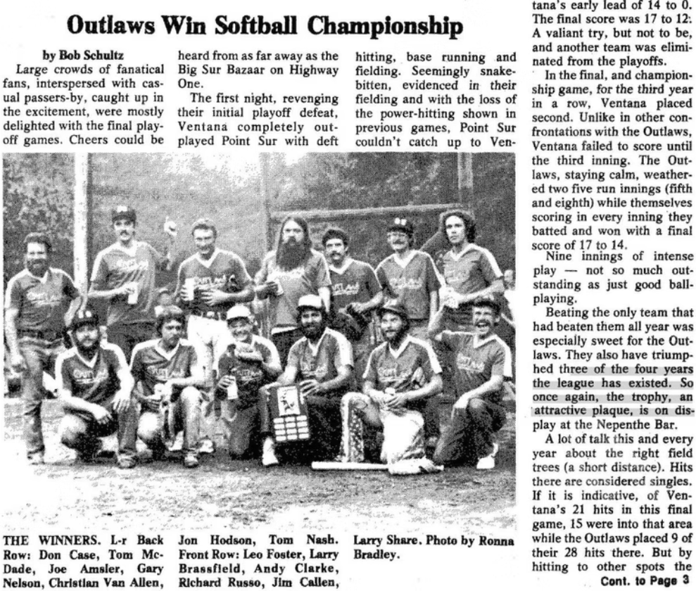 Group of baseball players posing with a trophy on a baseball field during daytime. They are wearing sports uniforms and caps, some are seated, and others are standing behind. A scoreboard and trees are visible in the background.