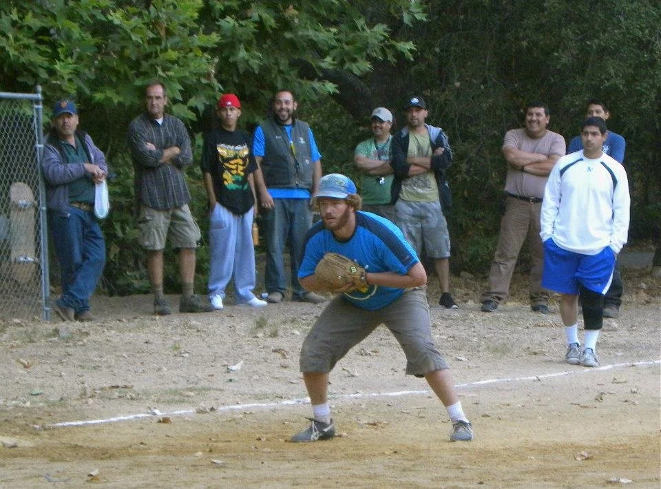 A man in a blue shirt and gray shorts playing baseball, standing on a dirt field with a glove ready. Several spectators are standing behind him, watching, with trees in the background.