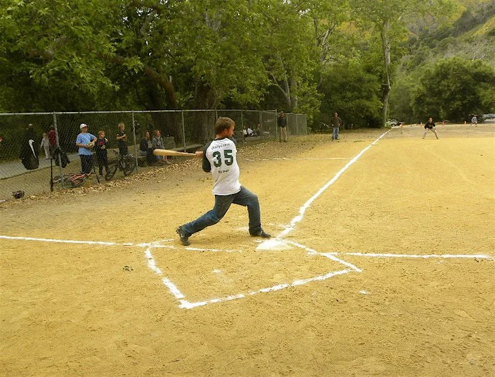 A young boy swinging a baseball bat at a baseball diamond during a game, with spectators and trees in the background.