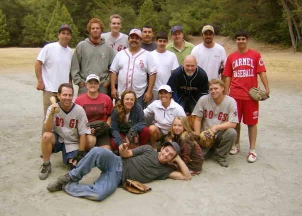 Group of people on a baseball field, posing for a photo during daytime, with trees in the background.