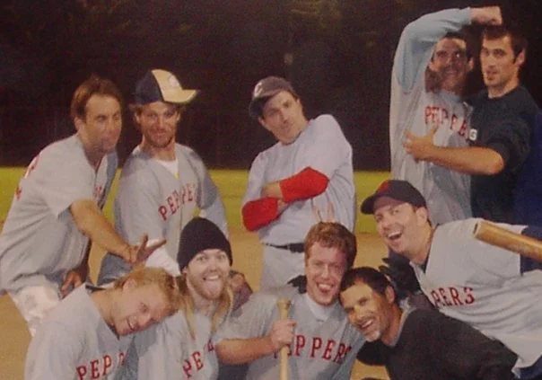 A group of baseball players celebrating on a field, some holding bats and wearing team uniforms, with a dark sky and trees in the background.