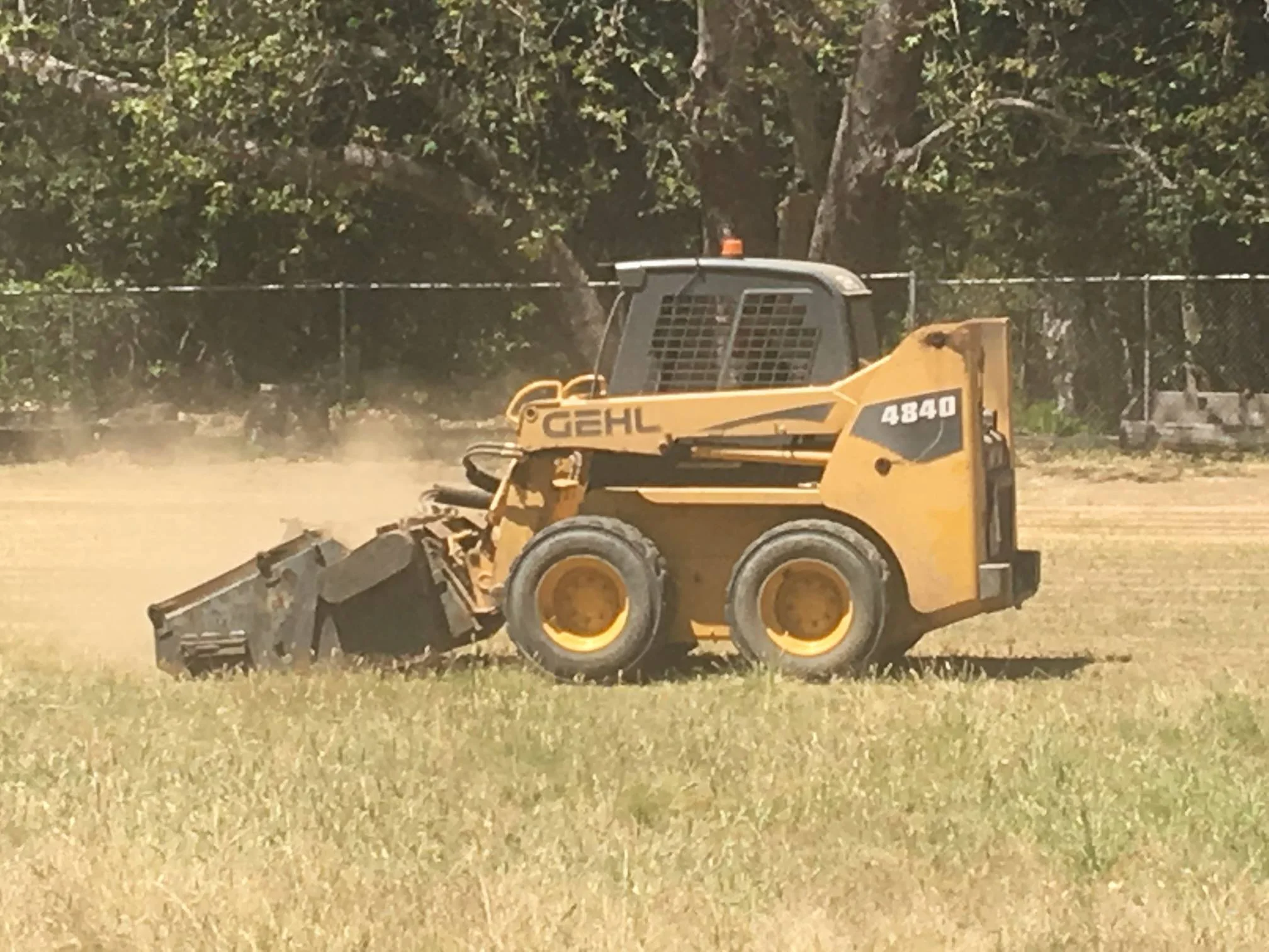 Yellow skid-steer loader with a front bucket attachment, labeled 'GHEL 4840', moving dirt or gravel on a grassy area.