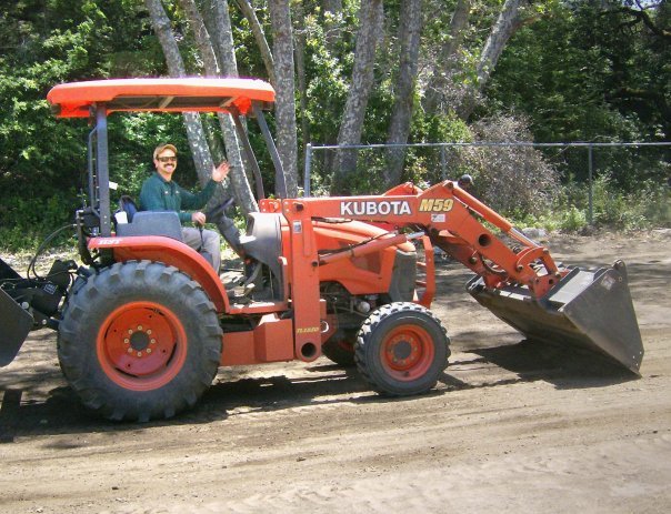 Person sitting on an orange Kubota M59 tractor with a front loader attachment, in an outdoor setting with trees in the background.