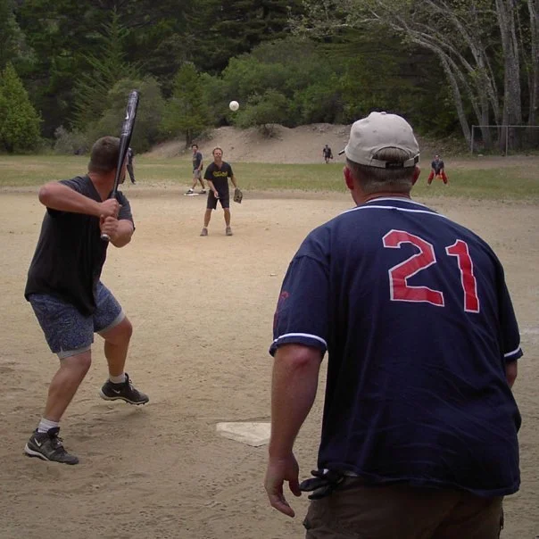 People playing baseball on a dirt field in a park with trees in the background.