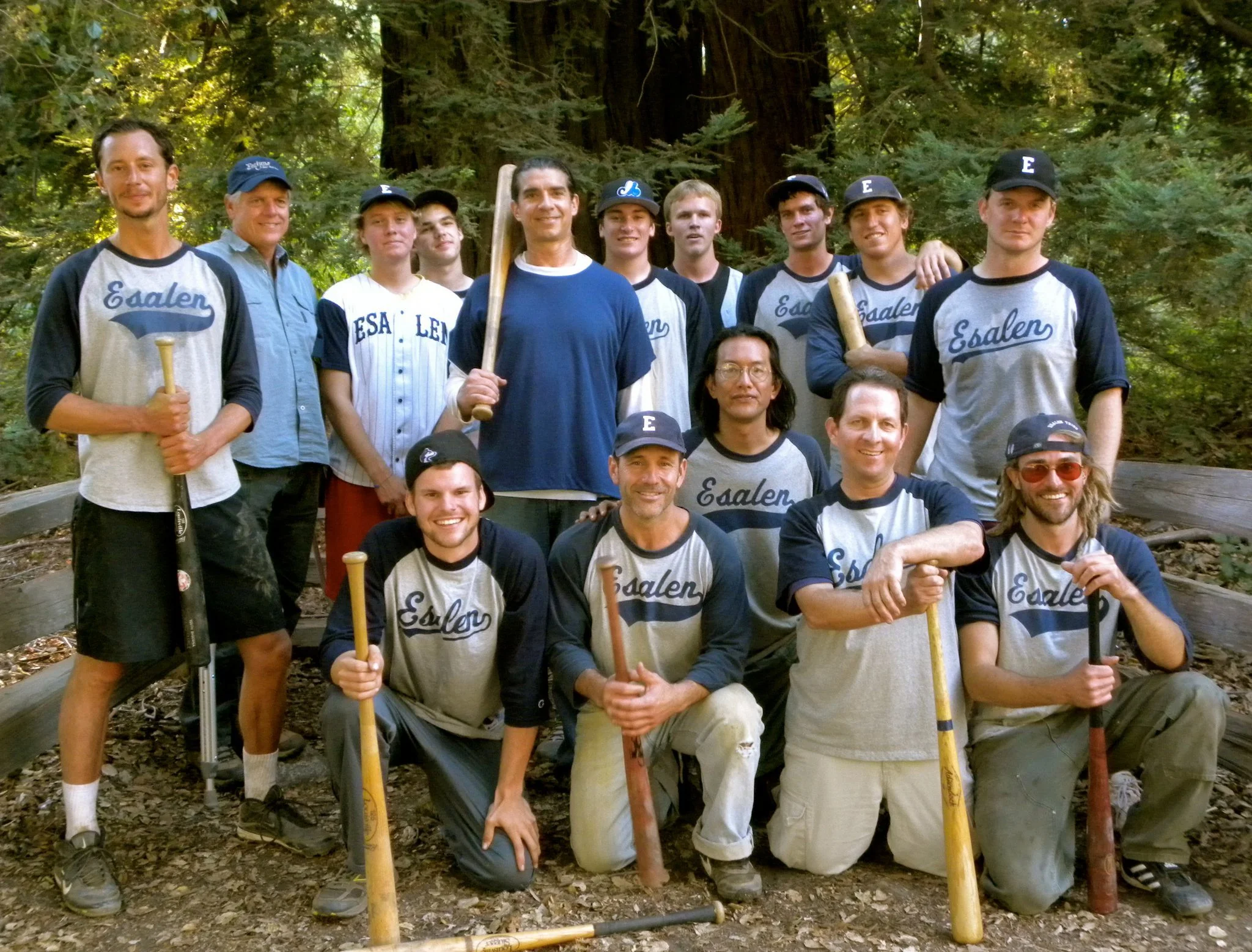 A group of young men and women in baseball uniforms, some holding bats, gathered outdoors in a forested area.