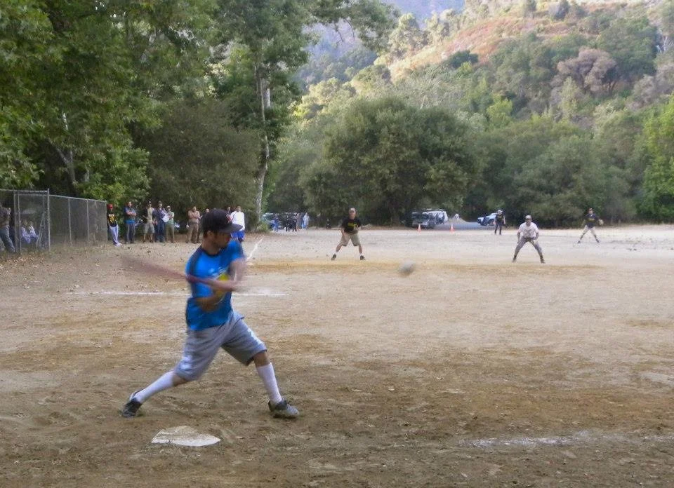 A young boy in a blue shirt and gray shorts swings a baseball bat at a pitched ball during a game, with other players and spectators in the background on a baseball field surrounded by trees.