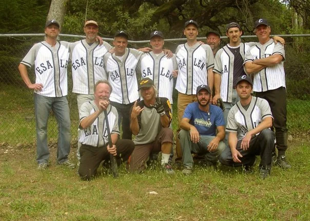 Group of ten men posing outdoors on grass, some wearing baseball jerseys with "ESA" and "LEA" on them, some with caps, in front of a chain-link fence and trees.