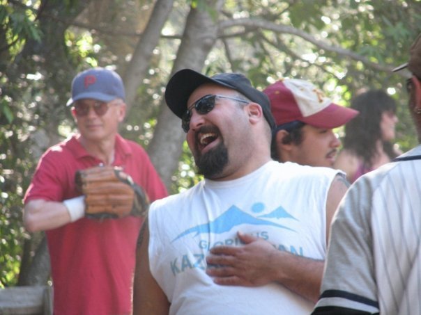 Group of men outdoors, one wearing a white sleeveless shirt, black cap, and sunglasses, laughing and singing, others in caps and casual attire, surrounded by trees.