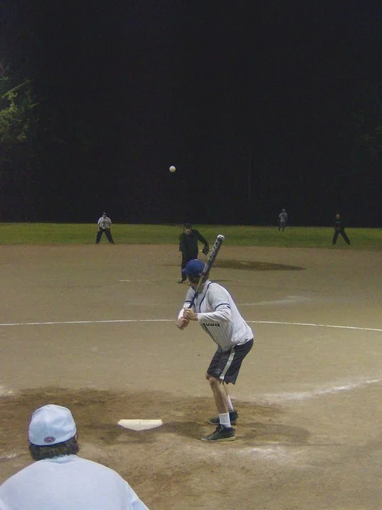 A baseball player prepares to pitch at night, with a baseball in mid-air, and other players on the field in the background.