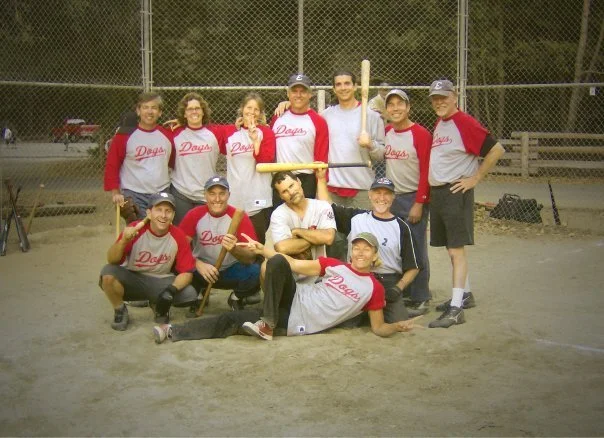A group of nine people on a baseball field, some wearing baseball uniforms and gloves, posing for a photo.