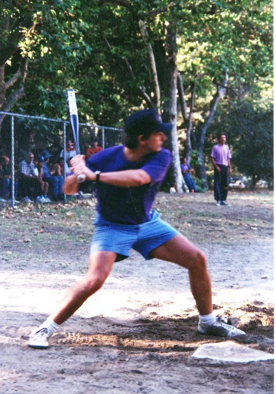 Person preparing to hit a baseball with a bat during a game at a park or field, with spectators and trees in the background.