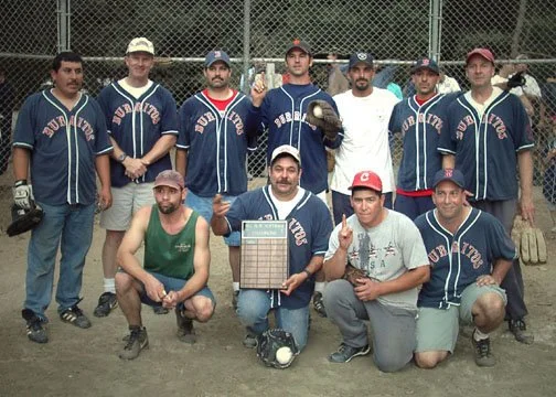 Group of men on a baseball field, some in jerseys, posing for a photo with one holding a plaque and others giving thumbs up.