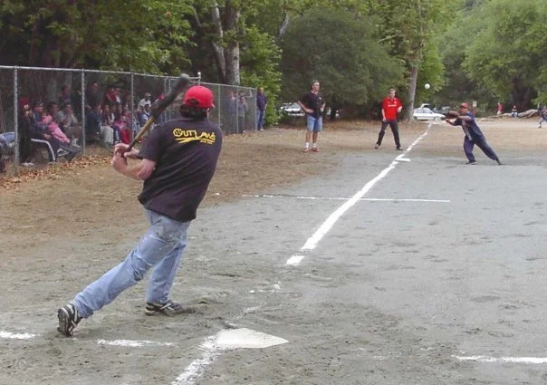 A baseball game taking place on a dirt field with a player swinging a bat, a pitcher throwing the ball, and spectators behind a chain-link fence.