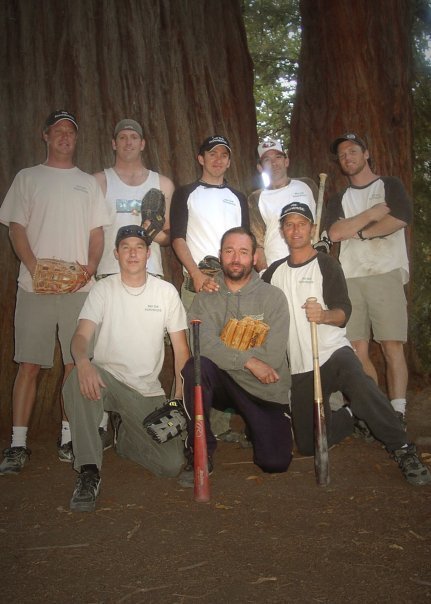 A group of eight men posing in front of large redwood trees, with some holding baseball or softball equipment, suggesting they are outdoors in a forested area.