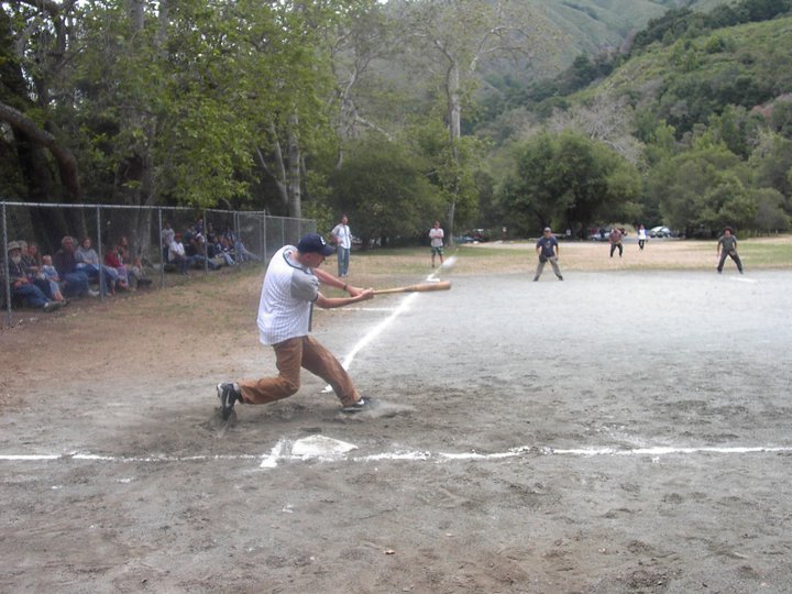 A person swinging a bat at a baseball game, with spectators sitting on the sidelines and other players on the field, outdoors surrounded by trees and hills.