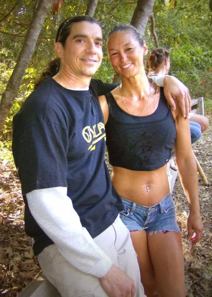 A man and woman smiling outdoors, with the woman having her arm around the man's shoulders and a young girl sitting on a bench in the background.