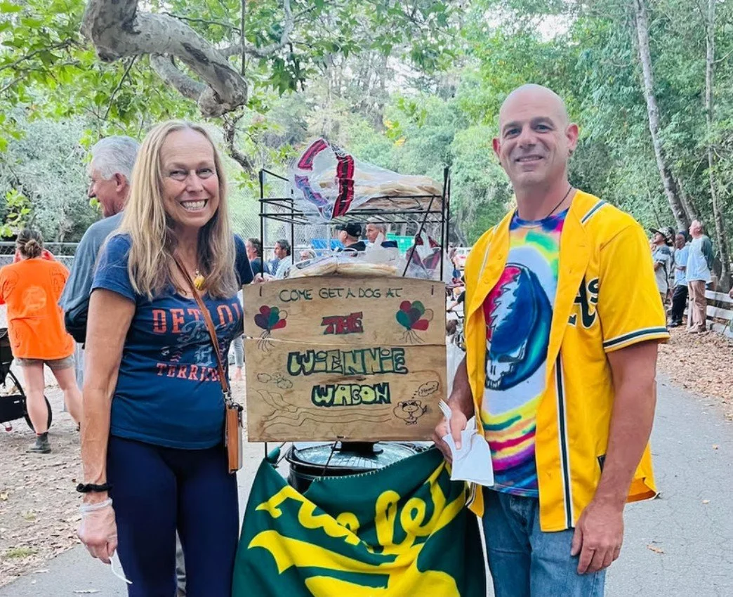 A smiling woman and man standing next to a wooden sign that reads 'Come get a dog at the WELCOME WAGON'. The sign is decorated with balloon and dog illustrations, and there is a yellow banner underneath. The scene is outdoors with trees and people in