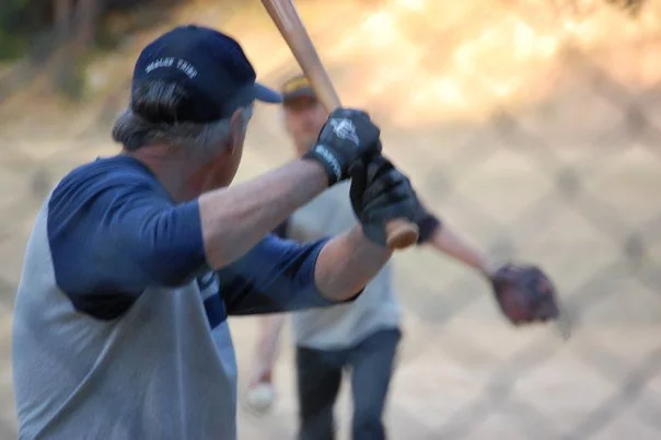 Older man swinging a bat, participating in a baseball or softball game outdoors.