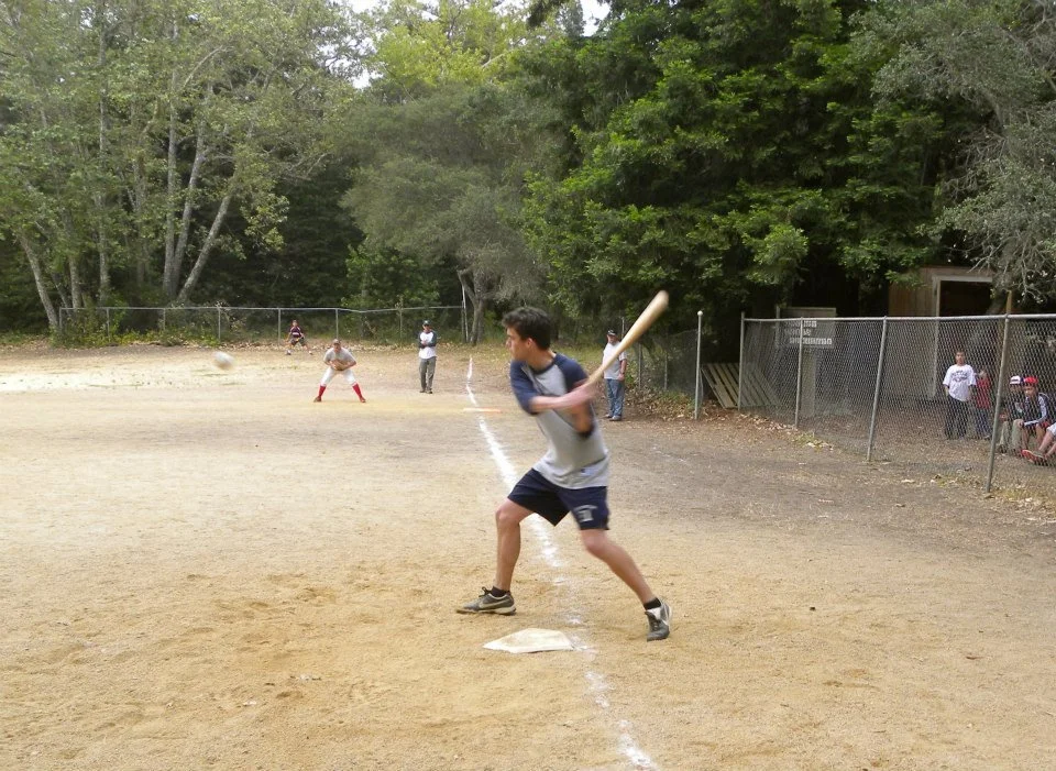 A boy batting in a baseball game on a dirt field with trees and spectators in the background.