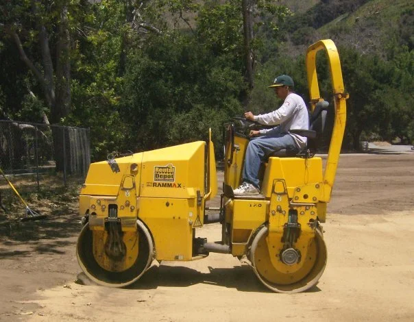 Man driving a yellow road roller compactor on a dirt path.