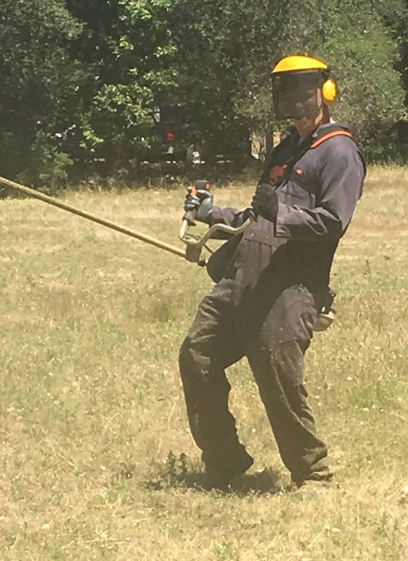 A person wearing a yellow safety helmet, goggles, and dark work clothes is holding a metal detector in an outdoor grassy area with trees in the background.