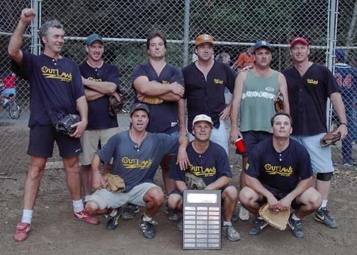 Group of seven men posing on a baseball field, some standing and some kneeling, with a large trophy or award placed on the ground in front of them, wearing matching team shirts with the word 'Outlaws'.