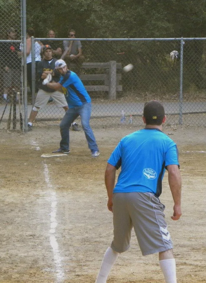 A baseball game at a park featuring a batter in a blue shirt and grey shorts, a catcher in a blue jersey catching the ball, and a pitcher in a blue shirt and grey shorts about to throw the ball. The game is happening on a dirt field with spectators b