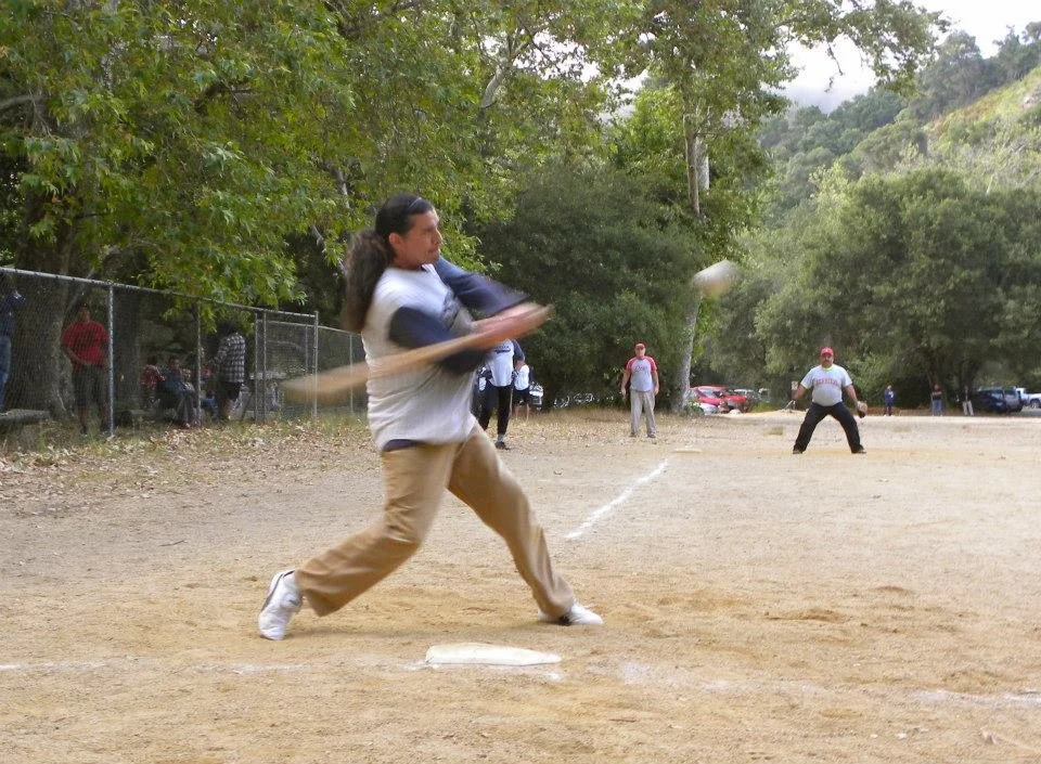 Person swinging a baseball bat at a baseball game on a dirt field with trees and spectators in the background.