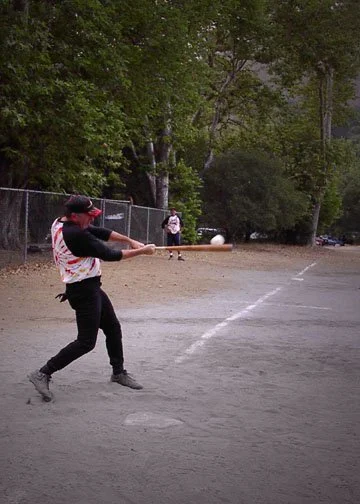 A person playing baseball on a dirt field, swinging a bat at a pitched ball, with trees and a chain-link fence in the background.