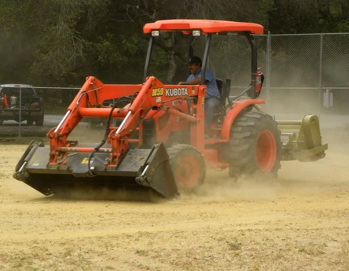 A man operating an orange Kubota tractor with a front loader attachment, working on a sandy or dirt field.