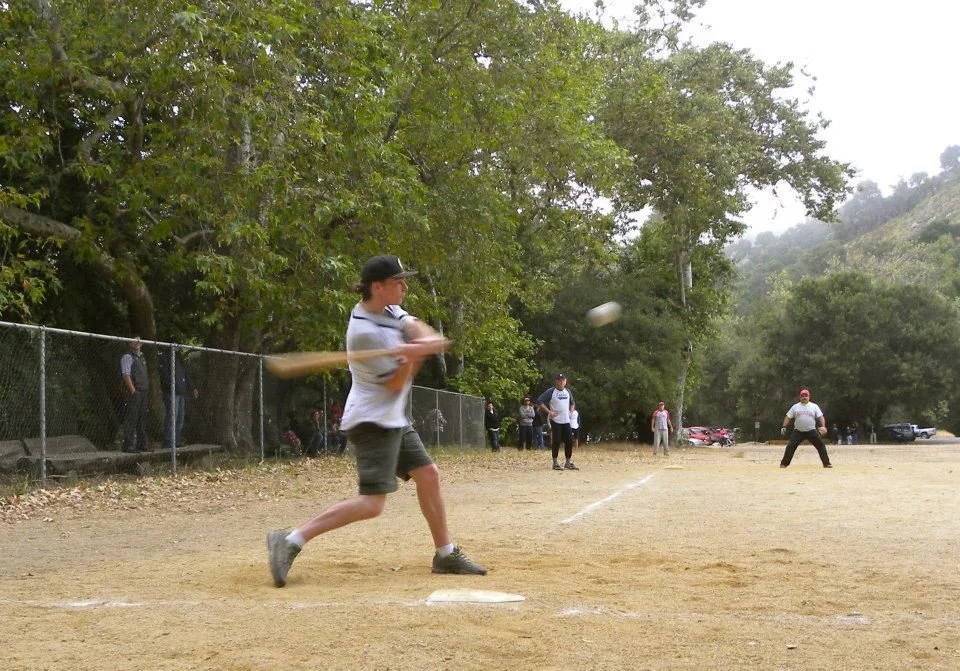 A person swinging a baseball bat at a ball on a baseball field, with several players and spectators in the background, trees around the field, and cars parked nearby.