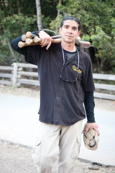 A man carrying a sledgehammer on his shoulder, wearing a black shirt and light-colored pants, standing outdoors near a wooden fence and trees.
