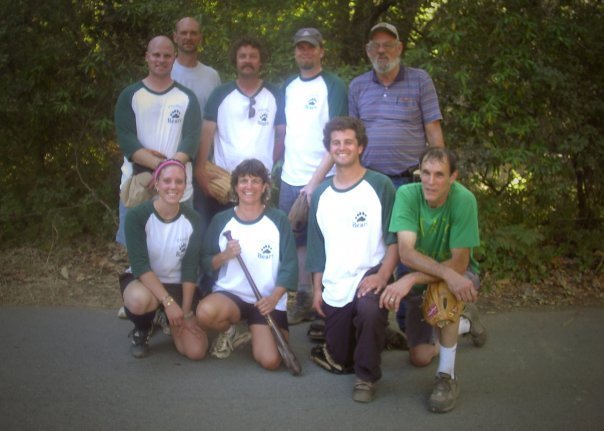 Group of nine people, including men and women, posing outdoors with greenery in the background, some wearing matching sports jerseys, one person holding a tennis racket, in a casual setting.