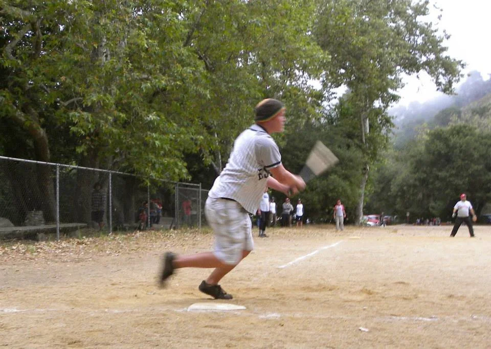 A young baseball player in uniform running towards a base during a game on a dirt field, with other players and spectators in the background.
