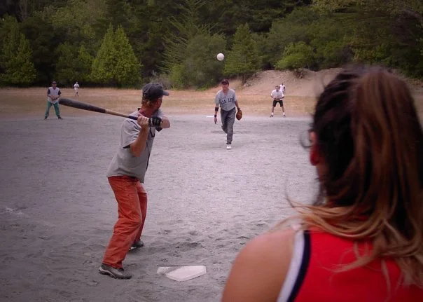 People playing baseball on a dusty field surrounded by trees.