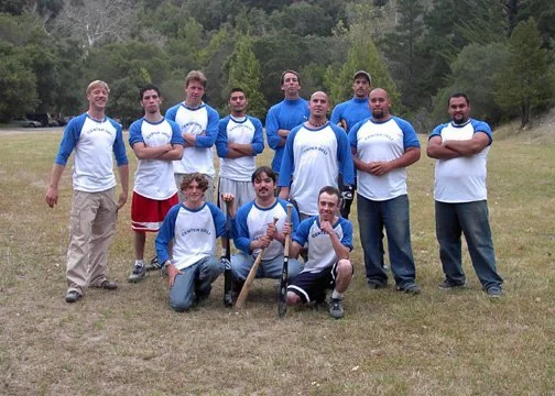 Group of nine young men standing and kneeling on grass field, some holding a baseball bat, outdoors with trees in background. All wearing matching blue and white long-sleeve shirts.