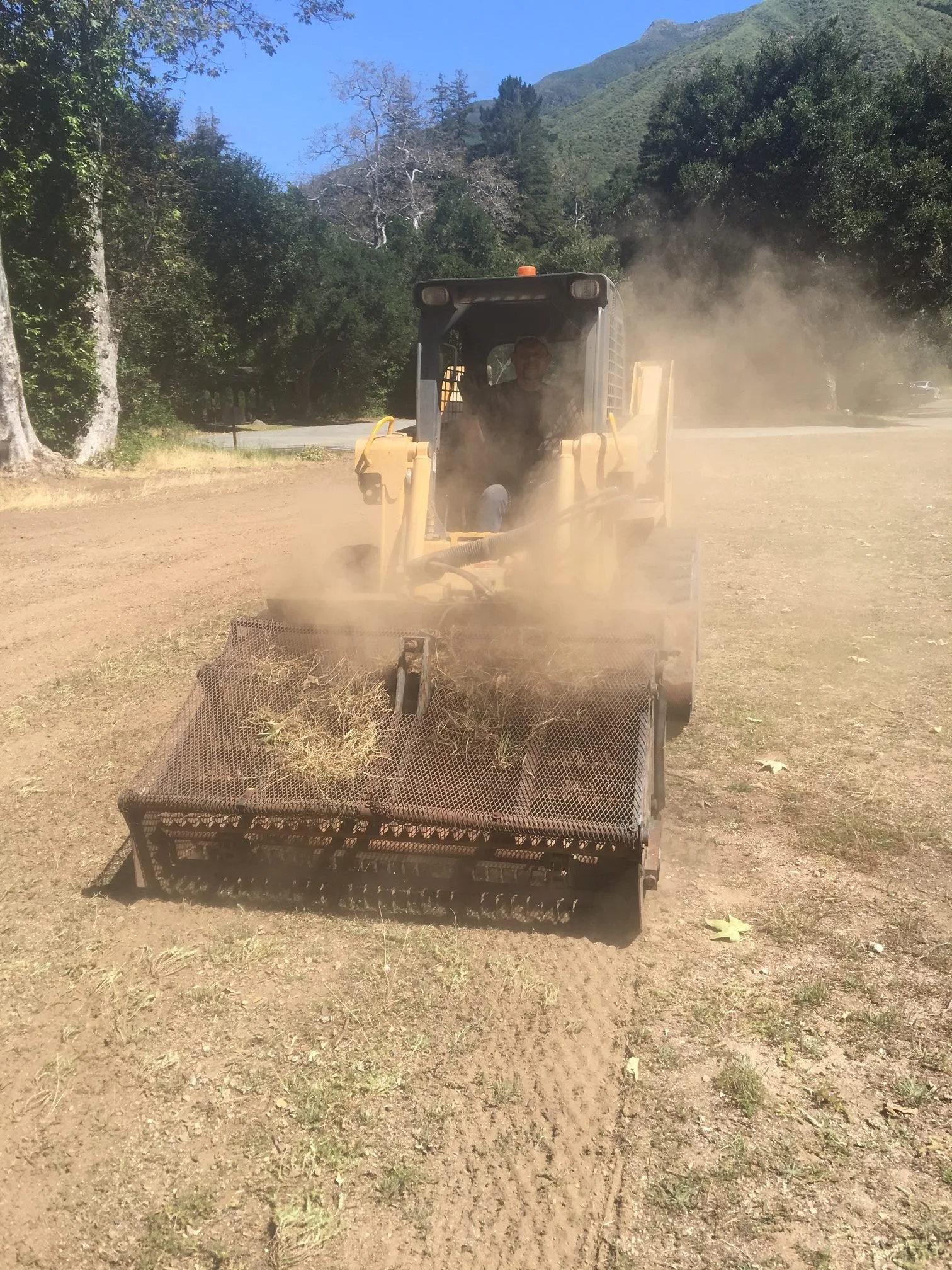 A person operating a piece of heavy machinery, possibly a wood chipper or mulcher, on a patch of dirt with a mountainous background and trees to the side, under a clear blue sky.