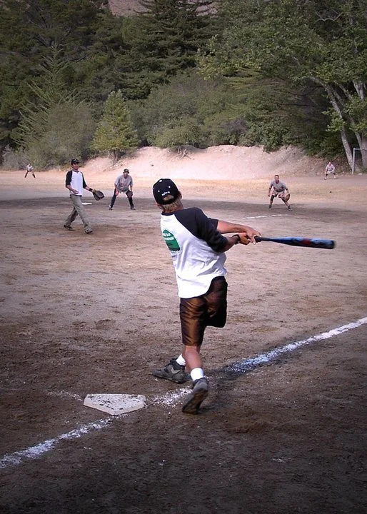 A young boy swinging a baseball bat at a baseball field with players in the outfield and trees in the background.