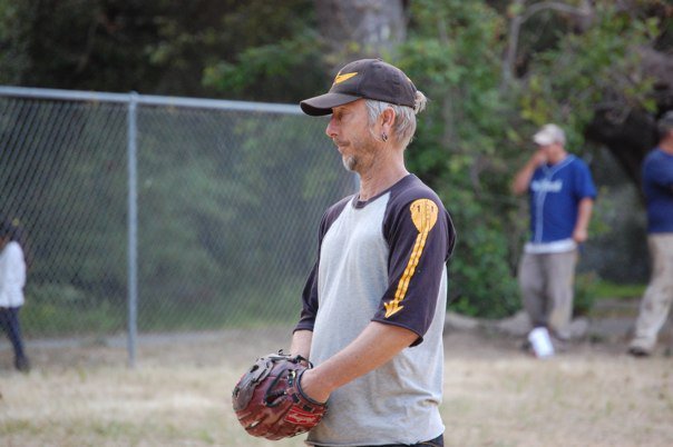 A man wearing a baseball cap and a gray and black baseball jersey with yellow accents, standing on a baseball field holding a baseball glove.