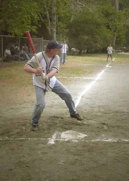 Teenager at bat during a baseball game on a dirt field, with spectators and other players in the background, surrounded by trees.