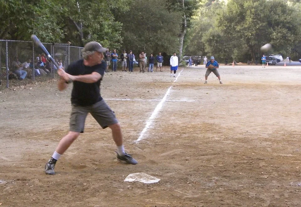 An older man in a black shirt and shorts preparing to hit a baseball with a bat on a baseball field, with people watching in the background.