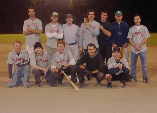 A group of eleven men posing on a baseball field, some in baseball uniforms and others in casual clothes, with a baseball bat on the ground in front of them.
