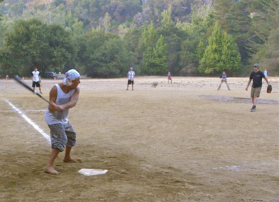 People playing baseball on a dirt field with trees in the background.