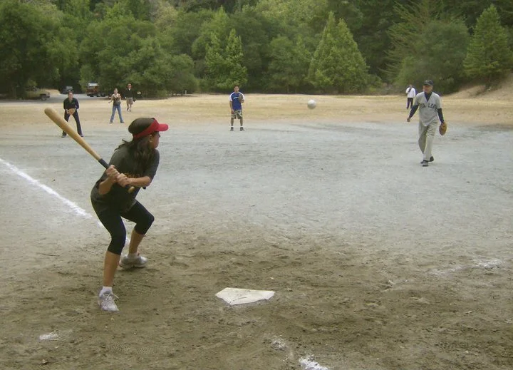 A girl playing softball at a baseball field with other players in the background and trees surrounding the field.