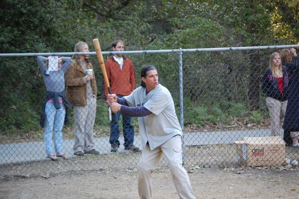 A man standing in a baseball batting stance holding a bat in front of a chain-link fence, with onlookers watching him, set in a park or backyard area.
